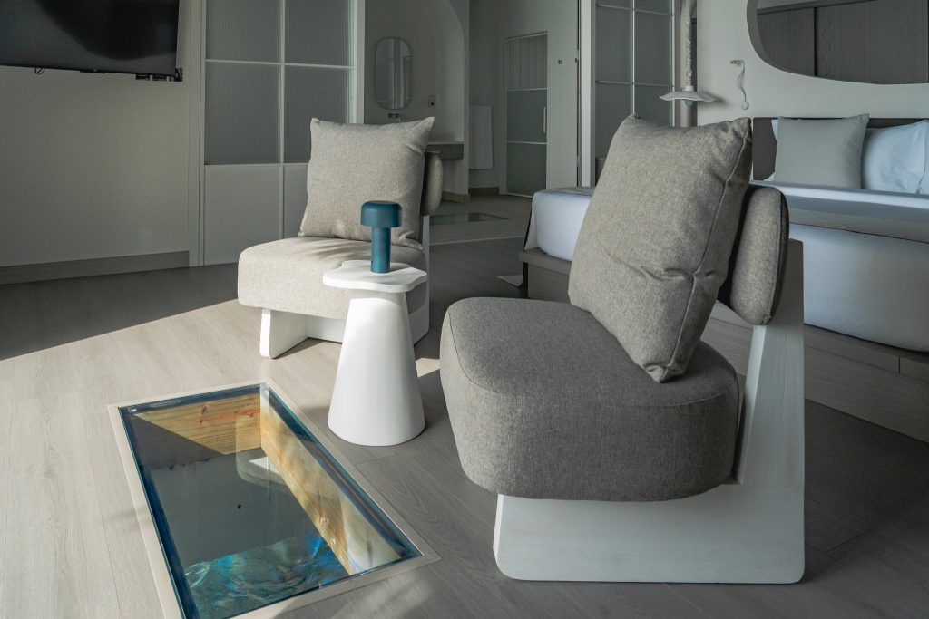 Modernes Hotelzimmer mit zwei grauen Stühlen, weißem Beistelltisch, Bodenfenster und hellem Holzboden im Sun Siyam Vilu Reef