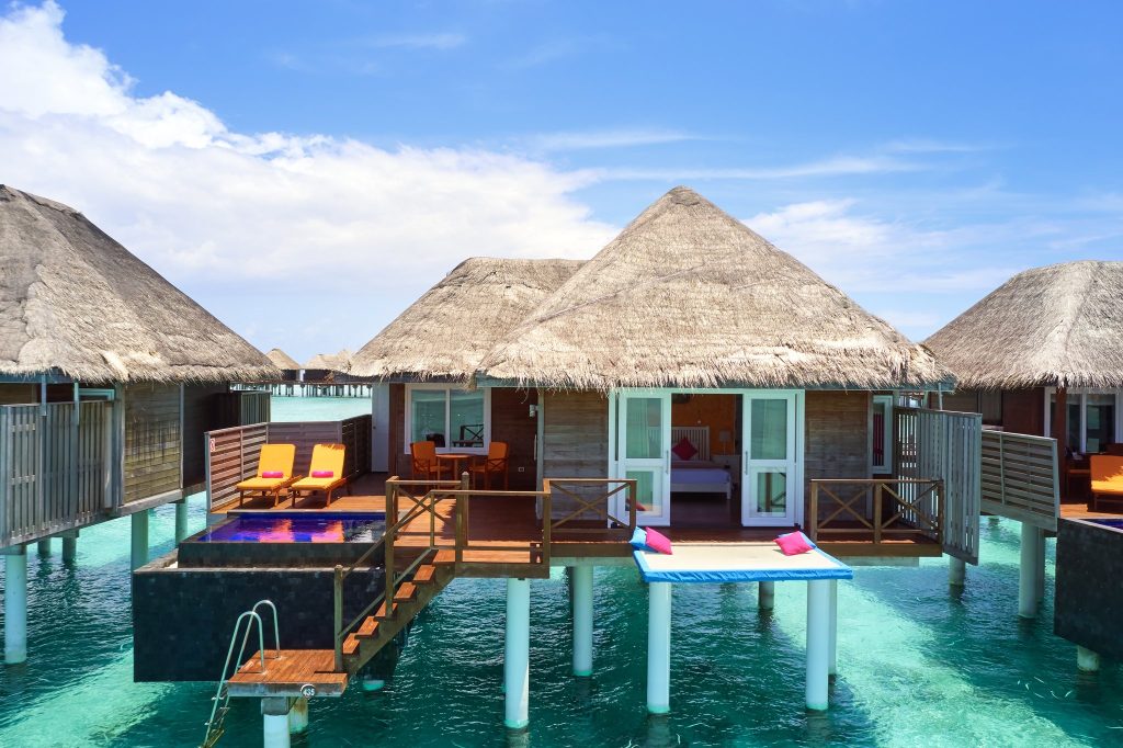 Water Villa mit Strohdach und Holzwänden auf Pfählen über türkisblauem Wasser mit privatem Pool und zwei Liegestühlen
